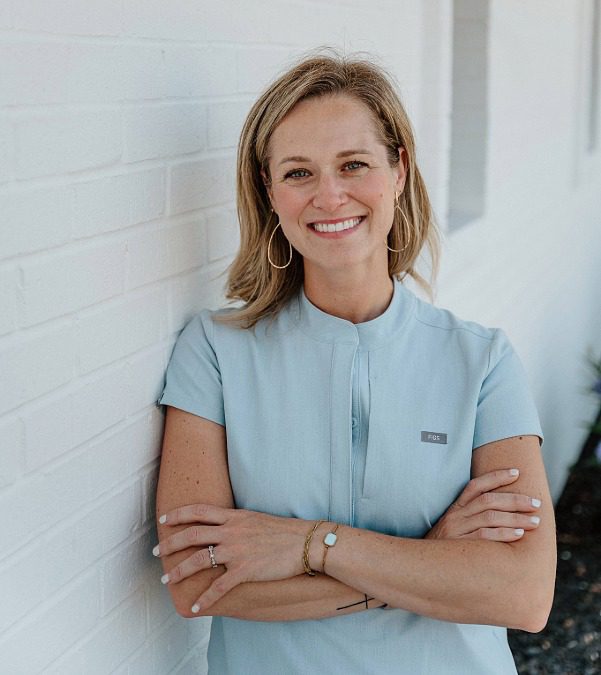 Female veterinarian leaning against a white brick wall wearing a light blue top
